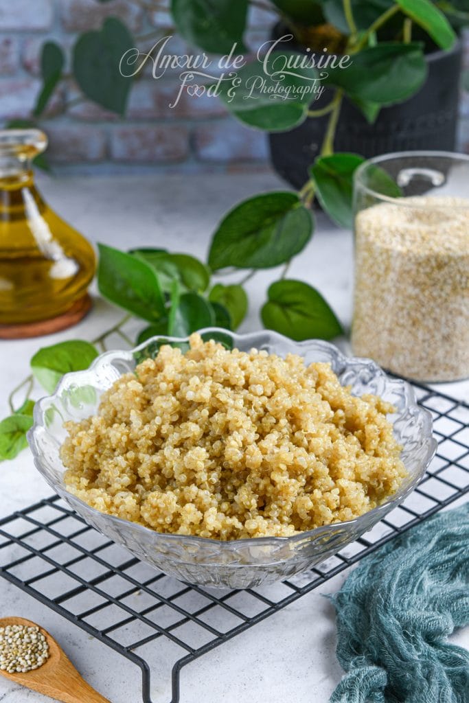 Bol de quinoa cuit, moelleux et doré, présenté dans un saladier en verre sur une grille de refroidissement, avec des graines de quinoa et une bouteille d’huile d’olive en arrière-plan.