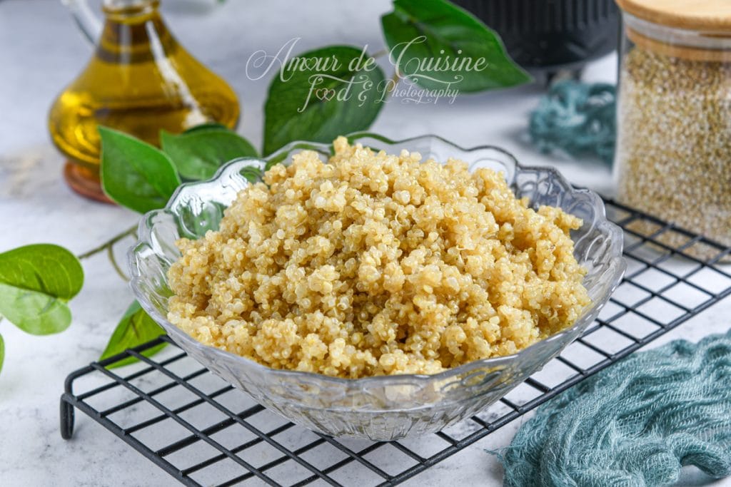 Bol de quinoa doré et moelleux servi dans un saladier en verre, posé sur une grille noire, avec une bouteille d’huile d’olive et un bocal de graines de quinoa en arrière-plan.