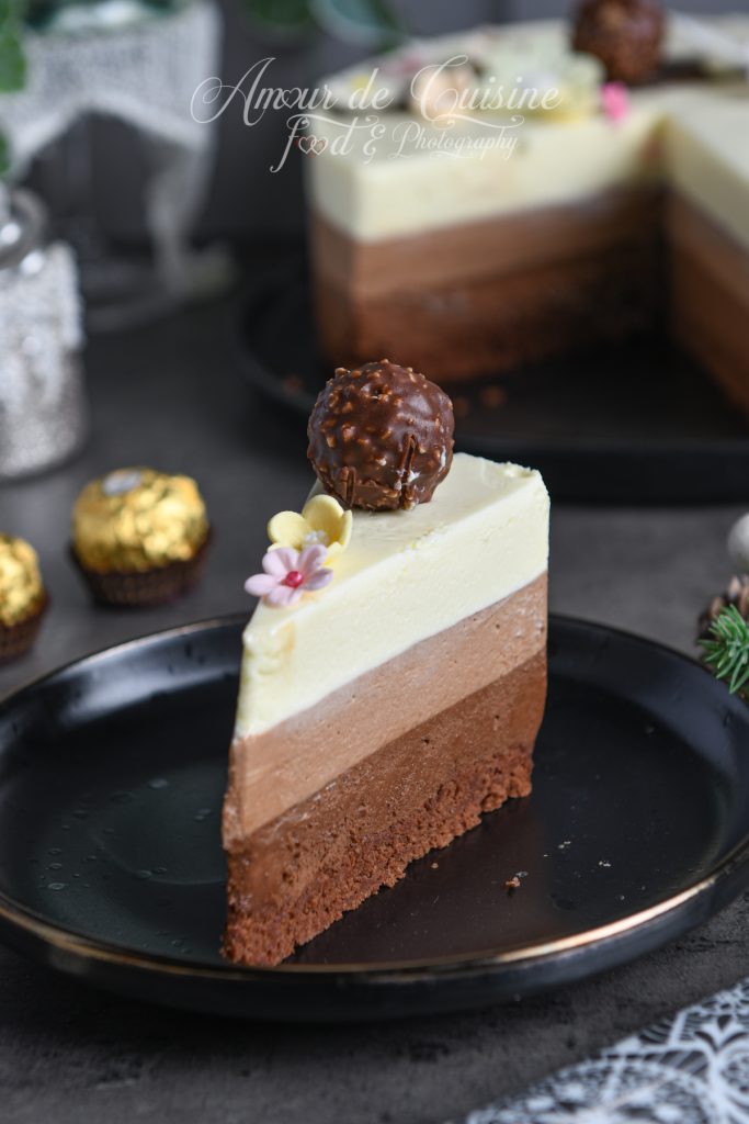 Part de gâteau entremets trois chocolats montrant les trois couches distinctes de mousse (chocolat noir, lait et blanc) sur une dacquoise cacao, décorée d’un Ferrero Rocher et de fleurs en sucre, servie sur une assiette noire.