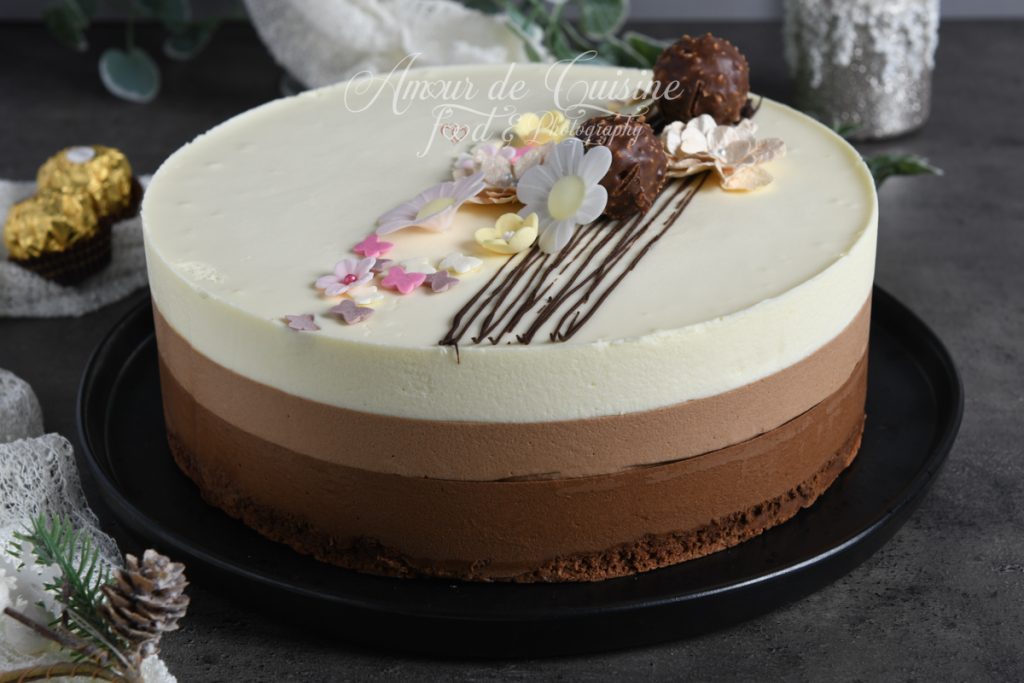 Entremets trois chocolats entier sur assiette noire, composé de trois couches de mousse (chocolat noir, lait, blanc) sur une base dacquoise, décoré de fleurs en sucre, lignes de chocolat et Ferrero Rocher.