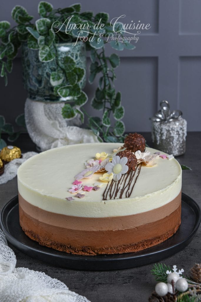 Entremets trois chocolats élégant, composé de trois couches de mousse (chocolat noir, lait et blanc) sur une base dacquoise cacao, décoré de fleurs en sucre et de Ferrero Rocher, présenté sur un plat noir.