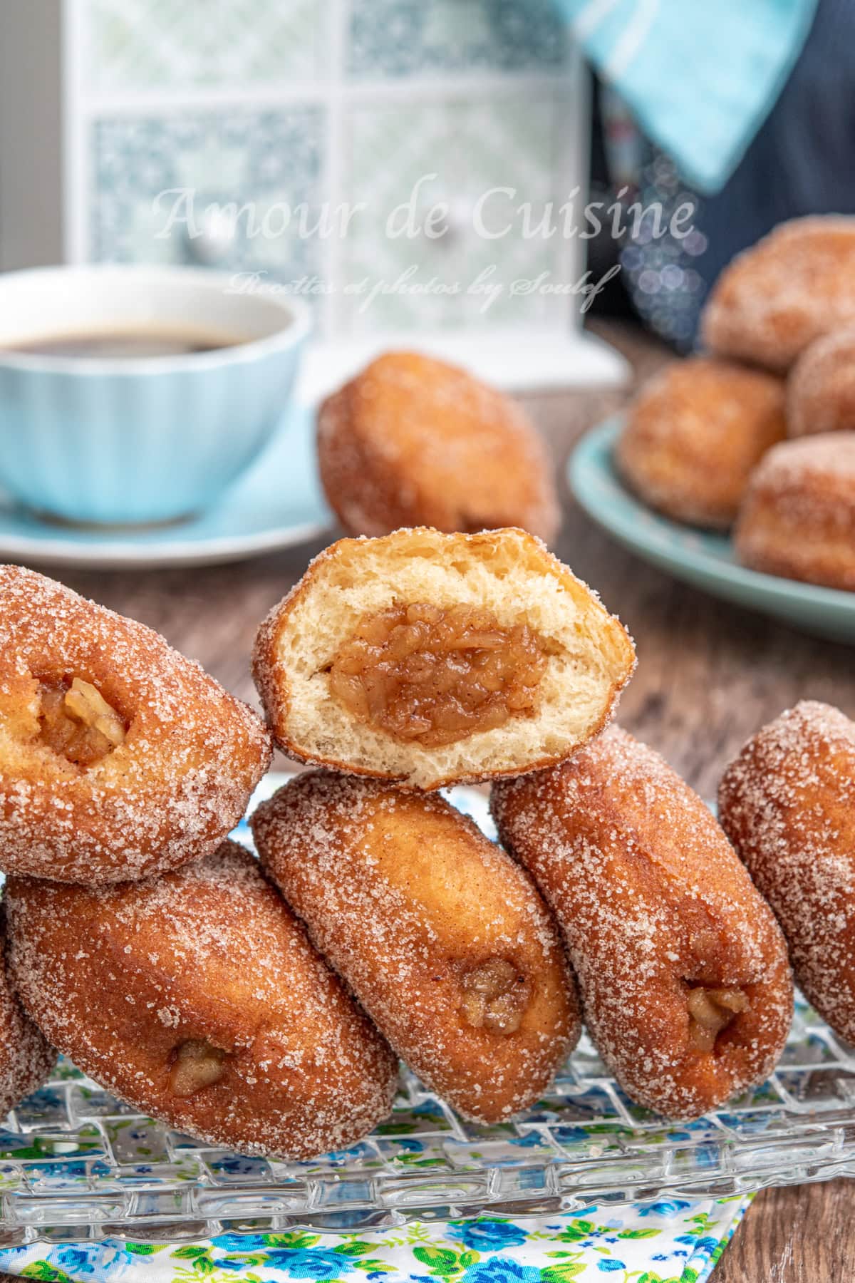 Close-up of sugar-coated beignets filled with apple compote, one cut open to reveal the gooey filling, served on a glass tray with a cup of coffee and more doughnuts in the background.