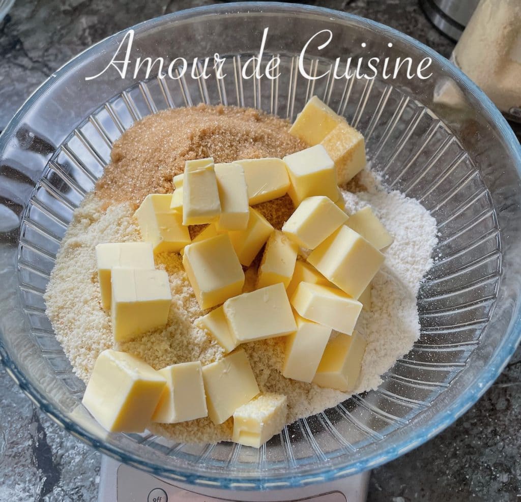 Saladier contenant des cubes de beurre, de la farine, de la poudre d’amandes et de la cassonade, prêts à être mélangés pour préparer une pâte à crumble maison.