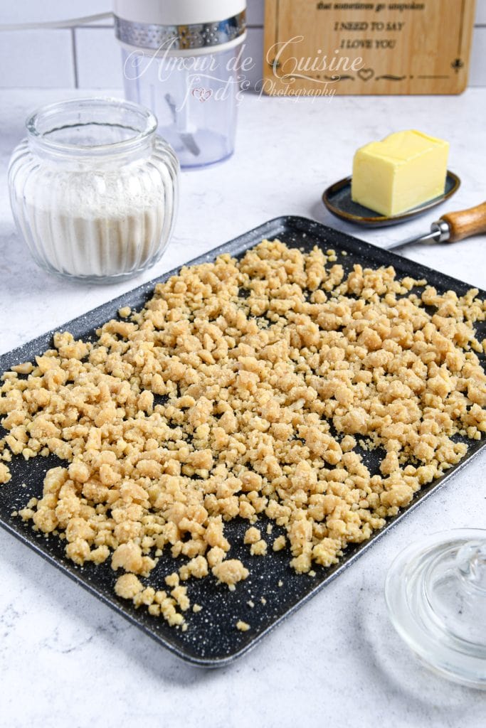 Plateau de pâte à crumble maison émiettée, prête à être cuite, posée sur un plan de travail avec un pot de farine, un bloc de beurre et un bol en verre en arrière-plan.