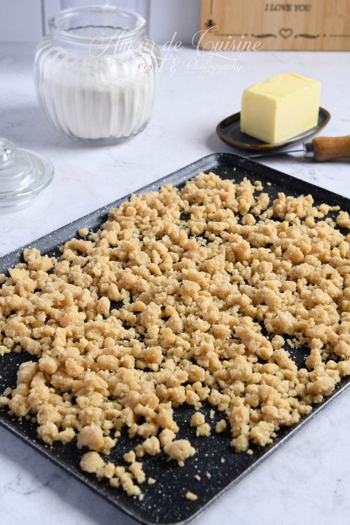 Plaque de cuisson recouverte de pâte à crumble émiettée, prête à être enfournée, avec en arrière-plan un pot de farine, un bloc de beurre et un plan de travail lumineux.