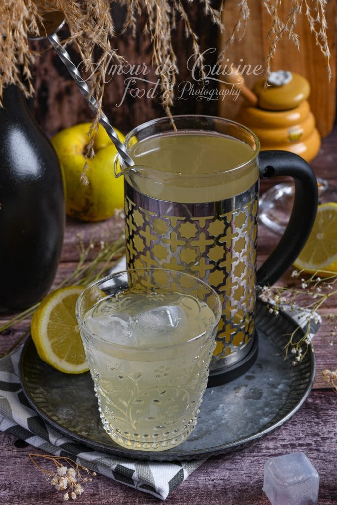Pichet de jus de coing maison décoratif, rempli d’un jus clair, avec un verre glacé au premier plan, entouré de tranches de citron, de coings jaunes et d’éléments rustiques sur une table en bois.