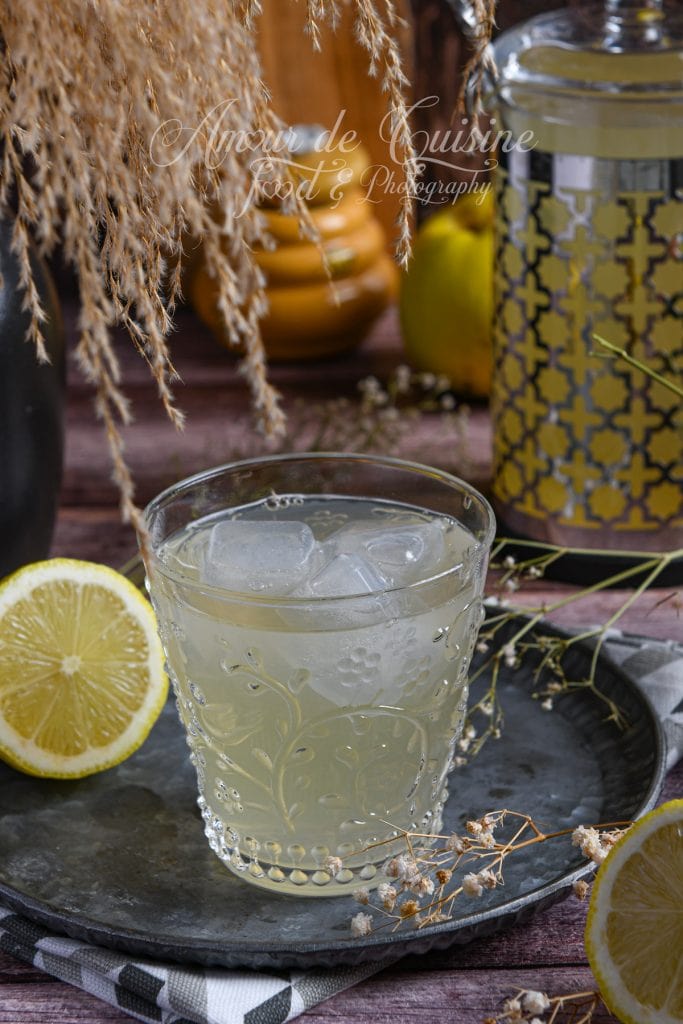 Verre de jus de coing maison rempli de glaçons, posé sur un plateau en métal, avec des moitiés de citron, un pot décoratif et des coings en arrière-plan dans une ambiance rustique et chaleureuse.