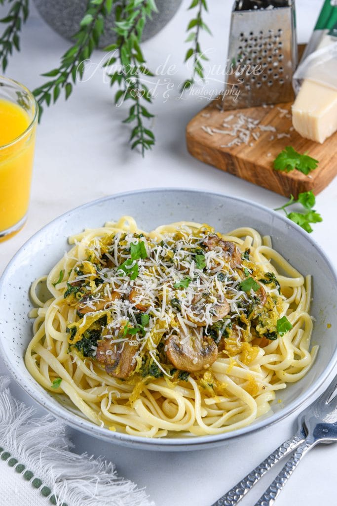 Assiette de spaghettis garnie de courge spaghetti, champignons sautés, épinards et parmesan râpé, avec un verre de jus et une râpe à fromage en arrière-plan.