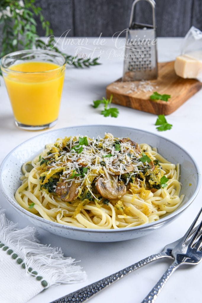 Assiette de spaghettis garnie de courge spaghetti, champignons sautés, épinards et parmesan râpé, servie avec un verre de jus d’orange et un fond de cuisine en arrière-plan.