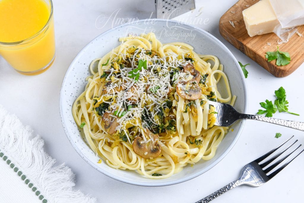 Assiette de pâtes garnie de courge spaghetti, champignons et épinards, parsemée de parmesan râpé, avec une fourchette enroulant les spaghettis et un verre de jus en arrière-plan.