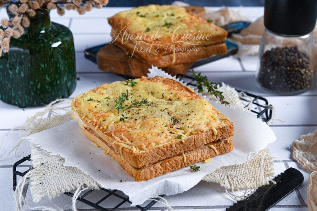Croque-monsieur à la mortadelle sans béchamel, gratiné et doré au fromage, servi entier sur une grille recouverte de papier cuisson, décoré de branches de thym frais, avec un moulin à poivre et un vase en arrière-plan