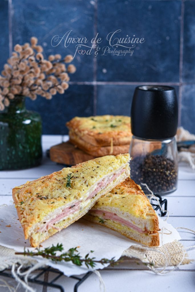 Croque monsieur à la mortadelle sans béchamel, bien doré et gratiné, coupé en deux et garni de fromage fondant et de fines tranches de charcuterie, présenté sur un papier cuisson avec une branche de thym, un moulin à poivre noir et une décoration florale en arrière-plan.