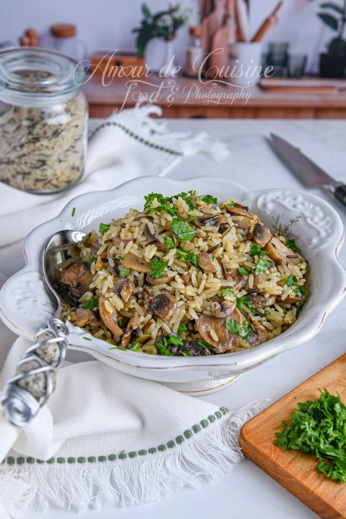 Bol de riz pilaf sauvage aux champignons, garni de persil frais, présenté dans un plat blanc élégant, avec un pot de riz sauvage, un couteau et un plan de travail de cuisine en arrière-plan.