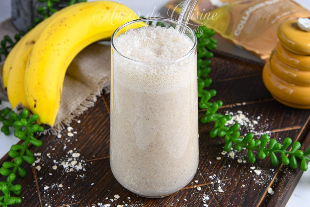 Verre de smoothie banane avoine crémeux posé sur un plateau en bois, entouré de bananes mûres, de flocons d’avoine et d’un pot de miel en arrière-plan.