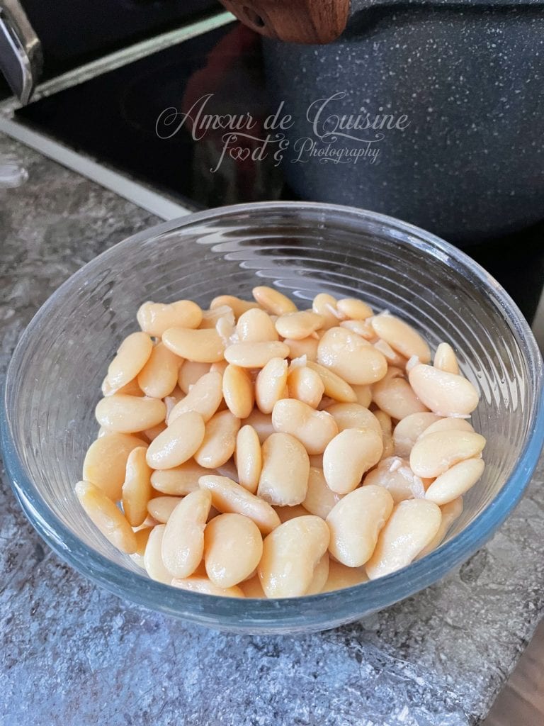 Bol en verre rempli de haricots beurre cuits et égouttés, posé sur un plan de travail de cuisine, avec une marmite en arrière-plan prête pour la préparation de la loubia.