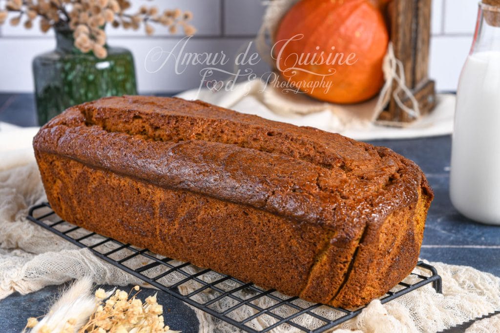 Whole extra moist pumpkin bread, golden and risen, placed on a cooling rack, with a bottle of milk, a pumpkin, and autumn-themed decor in the background.