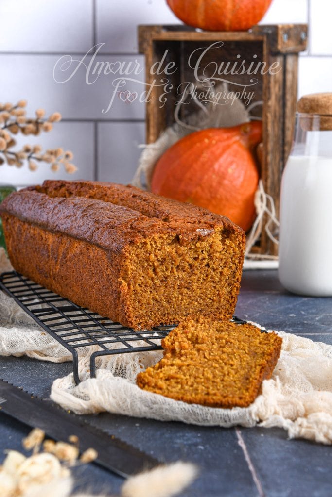 Extra moist pumpkin bread, sliced and placed on a cooling rack, with one slice in front; autumn decor with whole pumpkins, a bottle of milk, and rustic elements in the background.