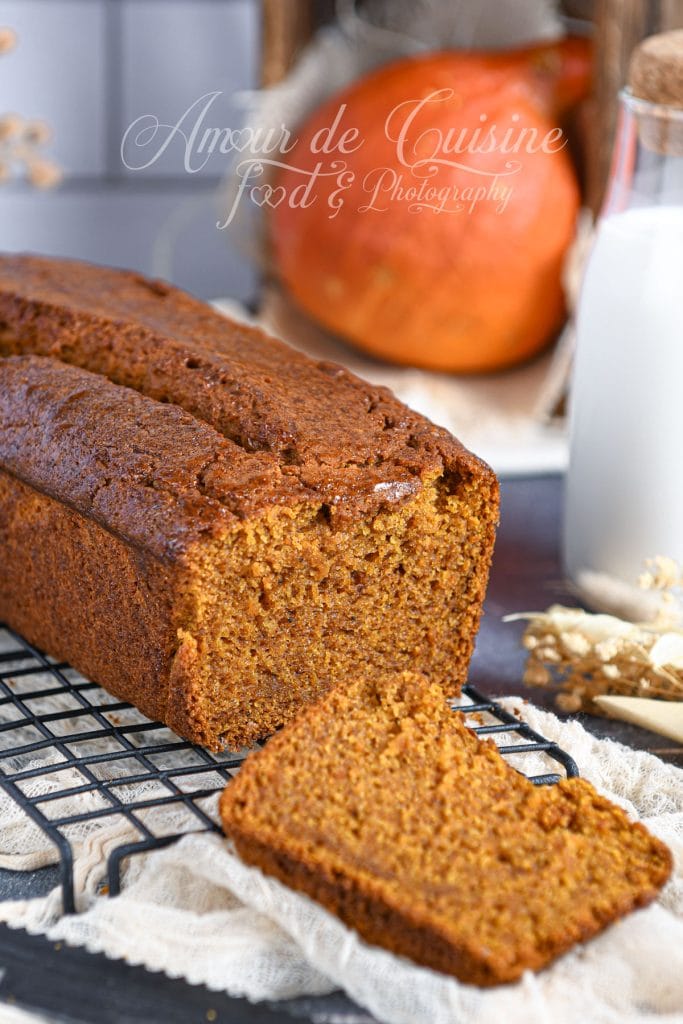 Extra-moist pumpkin bread, sliced, with one slice placed in front of the loaf on a cooling rack; autumn décor featuring a pumpkin and a bottle of milk in the background.
