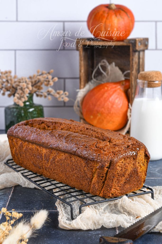 Whole extra-moist pumpkin bread, golden and glossy, placed on a cooling rack, with pumpkins, a bottle of milk, and autumn-themed decorations in the background.