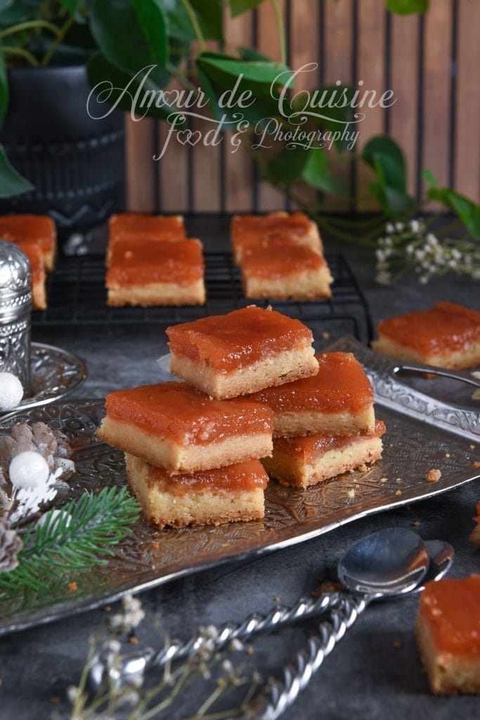 Carrés sablés à la pâte de coing, empilés sur un plateau argenté, avec une base sablée dorée et une garniture fondante à la pâte de coing, décor hivernal et ambiance chaleureuse en arrière-plan.