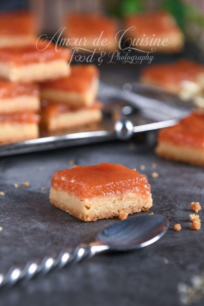 Un carré sablé à la pâte de coing en gros plan, posé sur une surface sombre, avec une base sablée dorée et une couche épaisse de pâte de coing, des miettes et une cuillère en premier plan.