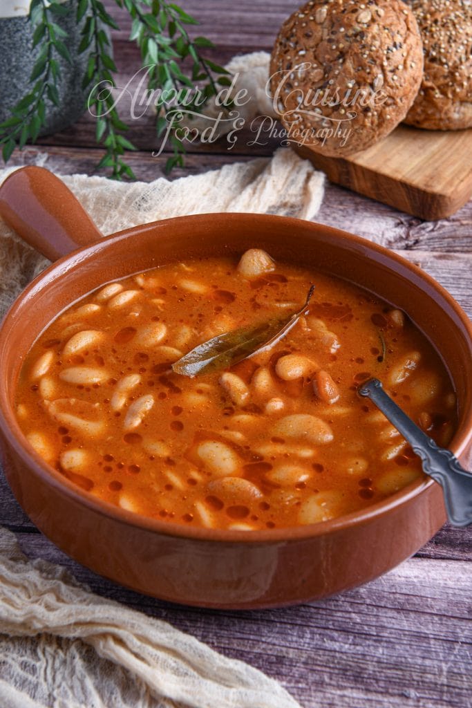 Cassoulet algérien aux haricots beurre servi dans une marmite en terre cuite, avec une sauce rouge onctueuse au cumin et une feuille de laurier, accompagné de petits pains aux céréales.