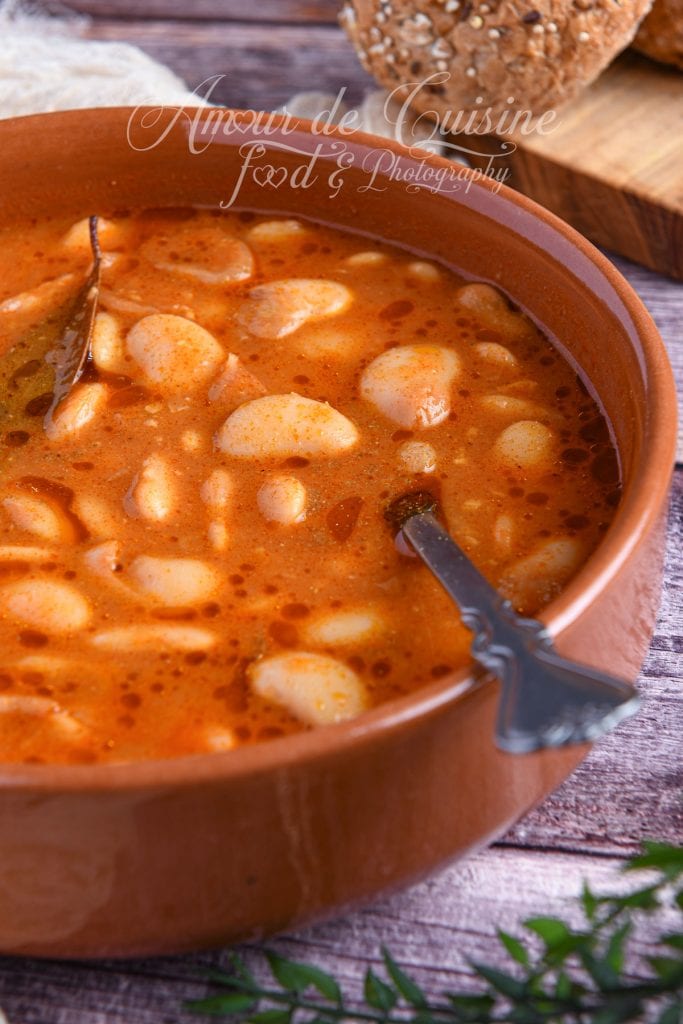 Gros plan d’un cassoulet algérien aux haricots beurre dans une marmite en terre cuite, avec une sauce rouge brillante au cumin et un brin de laurier, accompagné de pain aux céréales flou en arrière-plan.