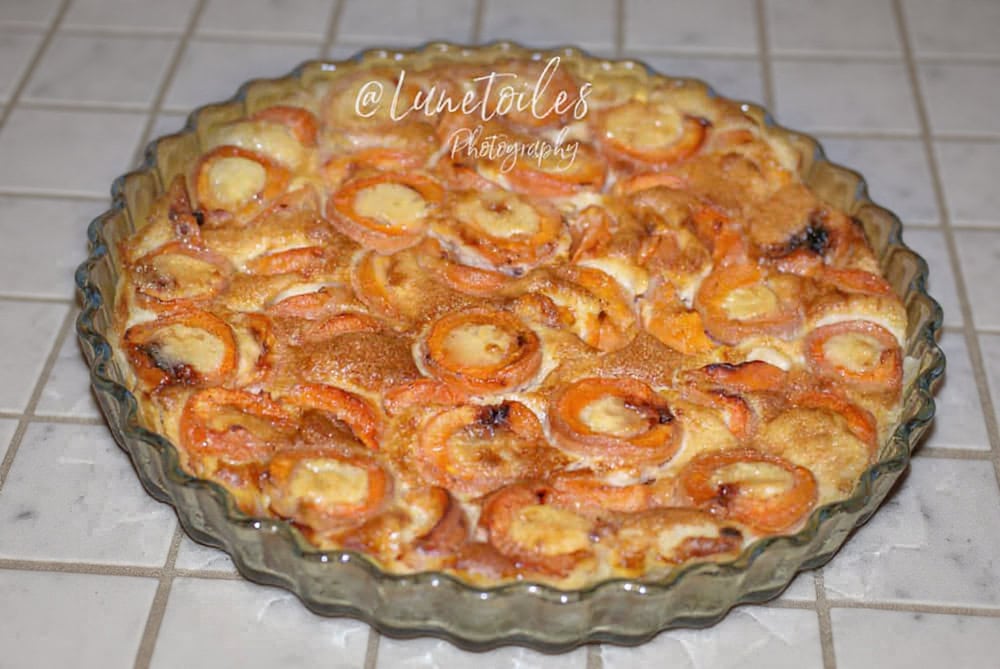 A freshly baked apricot clafoutis in a clear, fluted glass baking dish, set against a light grey background. The dessert features halved apricots nestled in a golden, custard-like batter with lightly browned edges and a glossy finish. The clafoutis appears rich and inviting, with the apricots slightly caramelized on top. The watermark "@Lunetoiles Photography" is visible across the center