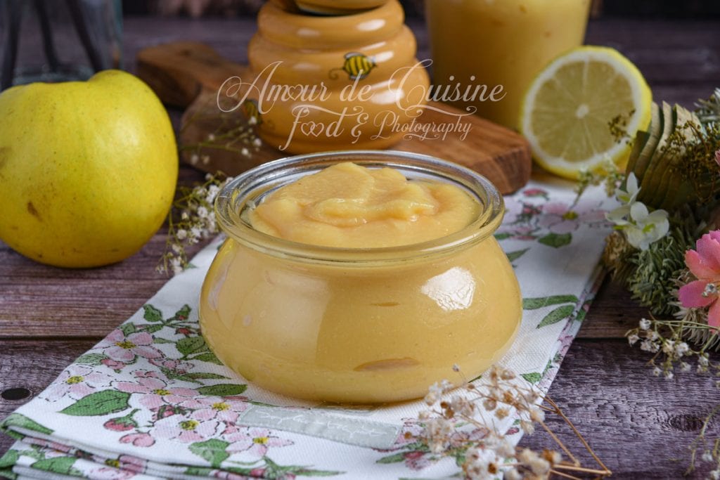 Honey quince sauce served in a small glass jar, surrounded by a fresh quince, a honey pot, a lemon half, and decorative flowers.
