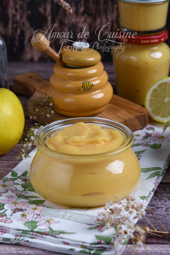 Sauce de coing au miel présentée dans un petit pot en verre, entourée d’un pot à miel, d’un bocal de sauce, d’un citron et d’une pomme, sur un torchon fleuri.