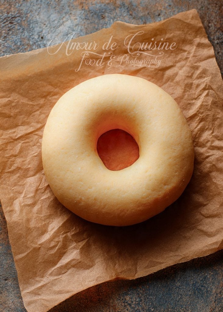 Uncooked donut dough shaped into a perfect ring, resting on a sheet of crinkled brown parchment paper, showing its smooth, soft texture before frying or baking.