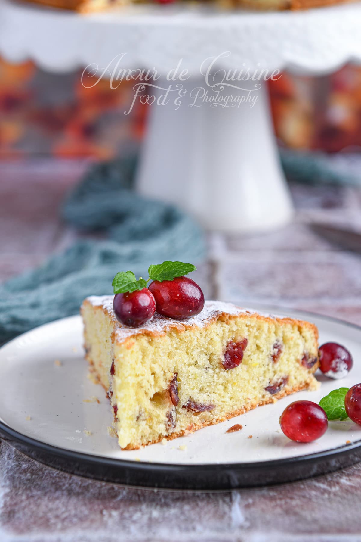 Slice of moist cranberry cake on a white plate, topped with fresh cranberries and a mint leaf, with a blurred cake stand and warm autumn tones in the background.