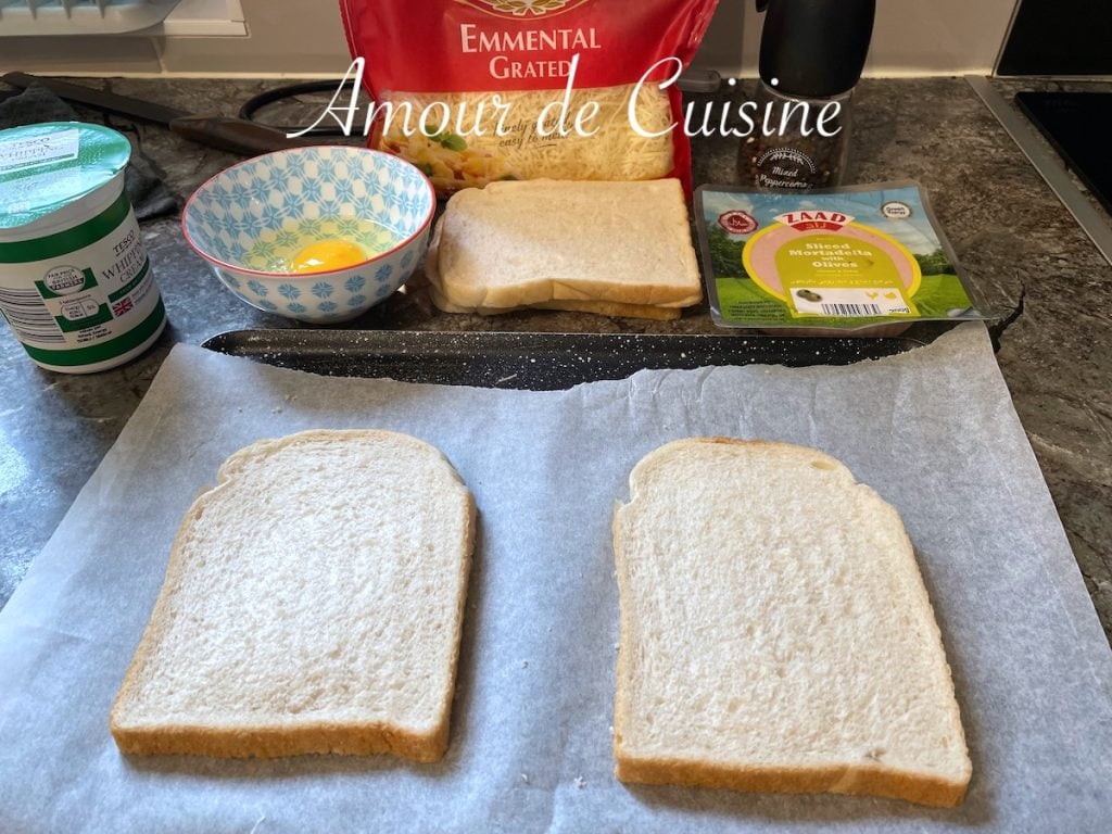 Ingrédients pour croque monsieur à la mortadelle sans béchamel : tranches de pain de mie disposées sur une plaque recouverte de papier cuisson, avec un bol contenant un œuf, un sachet de fromage râpé, de la mortadelle, de la crème fraîche, et un moulin à poivre posés à l’arrière sur le plan de travail.