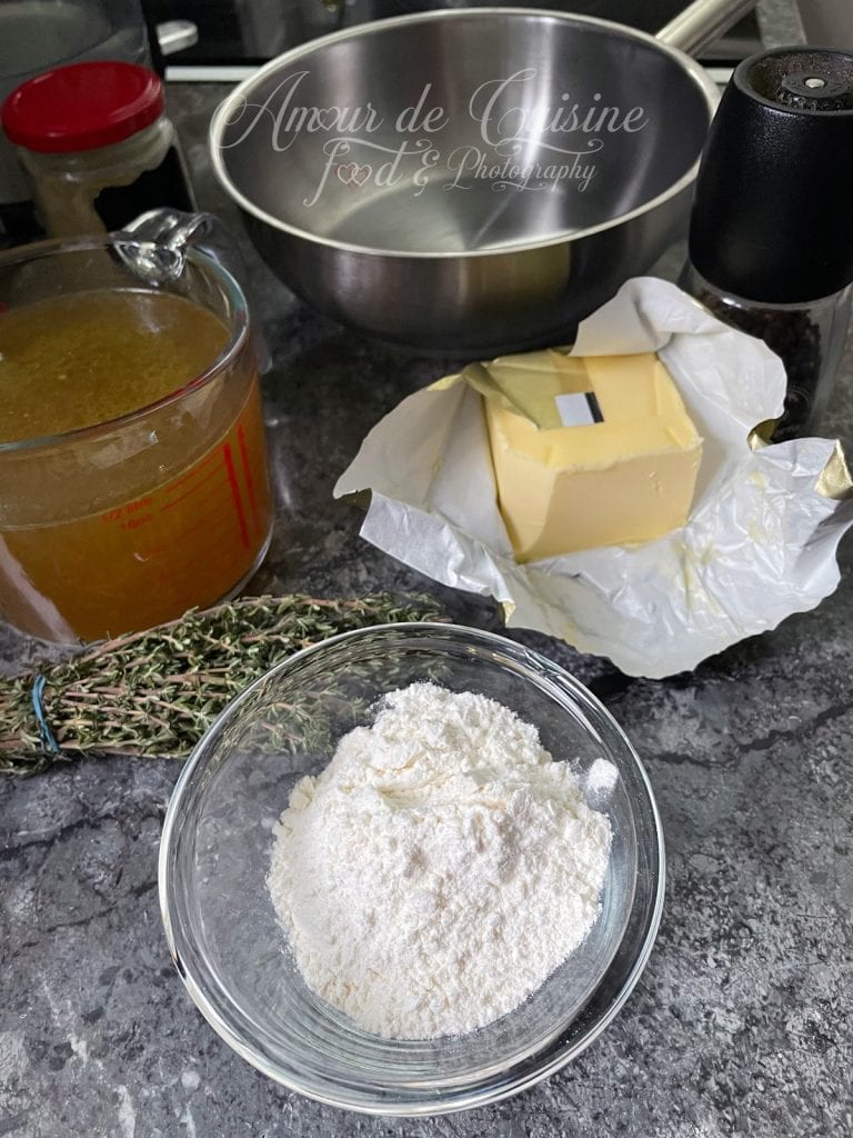 Ingrédients pour préparer une sauce brune disposés sur un plan de travail : un bol de farine blanche, un bouquet de thym frais, un pichet de bouillon chaud, une grande casserole en inox vide, un morceau de beurre sur son papier d’emballage et un moulin à poivre noir.