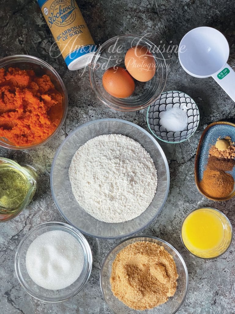Ingredients for pumpkin bread: flour, pumpkin purée, eggs, white sugar, brown sugar, oil, orange juice, baking soda, salt, and a blend of spices arranged on a stone countertop.