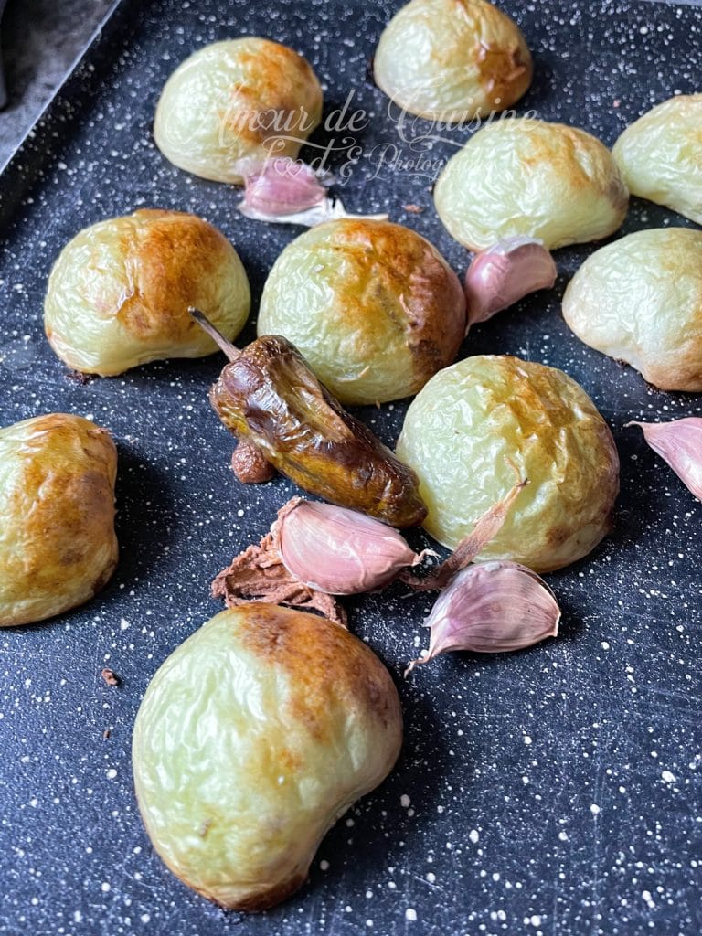 Tomatillos rôtis sur une plaque de cuisson noire, leur peau légèrement brunie et cloquée, accompagnés de gousses d’ail rôties encore en peau et d’un piment grillé. L’ensemble montre la cuisson uniforme et la texture fondante typique des ingrédients destinés à une salsa verde maison.