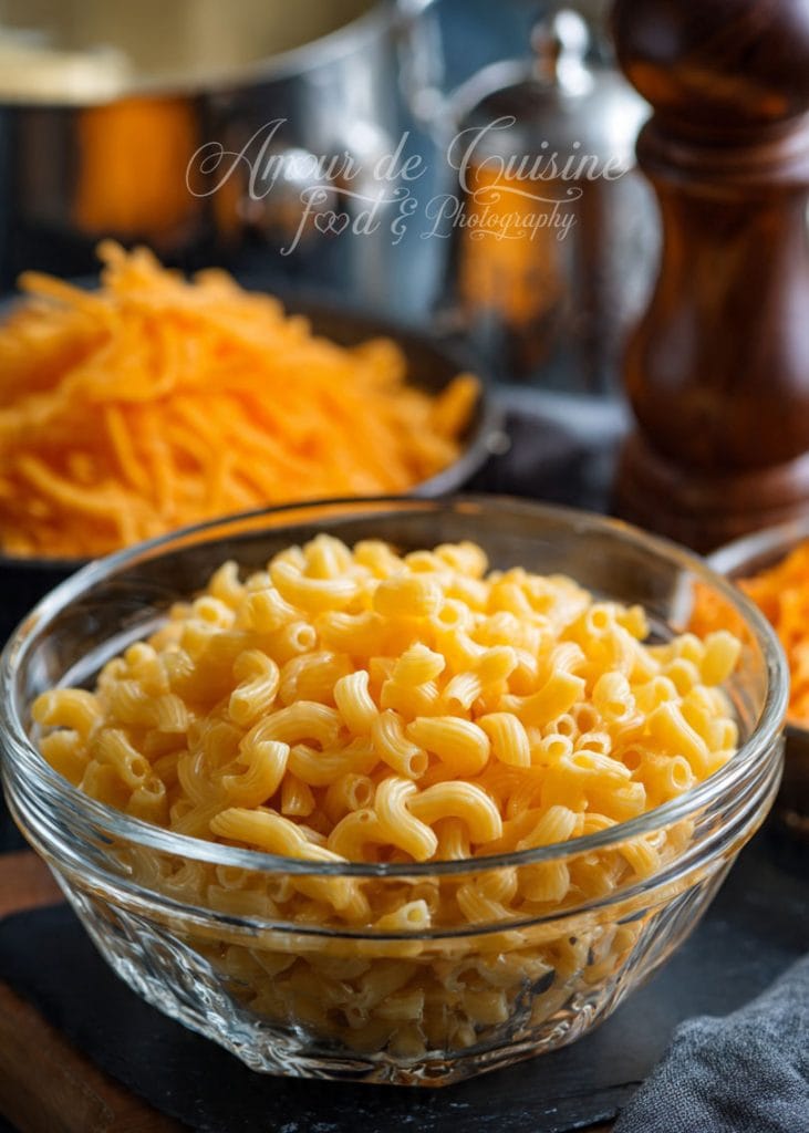 Bol en verre rempli de macaronis cuits, posé devant des bols de cheddar râpé, prêt pour la préparation d’un mac and cheese crémeux, avec ustensiles de cuisine en arrière-plan.