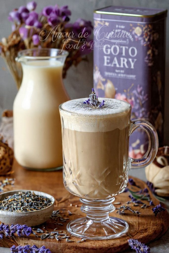 A creamy London Fog latte served in an elegant glass mug, topped with frothy milk and a lavender sprig, placed beside a small bowl of dried lavender, a jug of milk, and a decorative Earl Grey tea tin with purple floral accents.