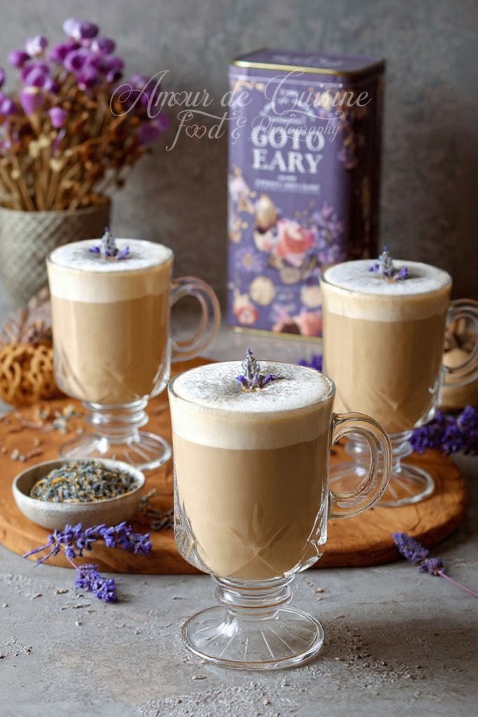 Three creamy London Fog lattes served in elegant glass mugs, each topped with frothy milk and a small lavender sprig, displayed on a rustic board with loose lavender, a bowl of dried flowers, and a decorative Earl Grey tea tin in the background.