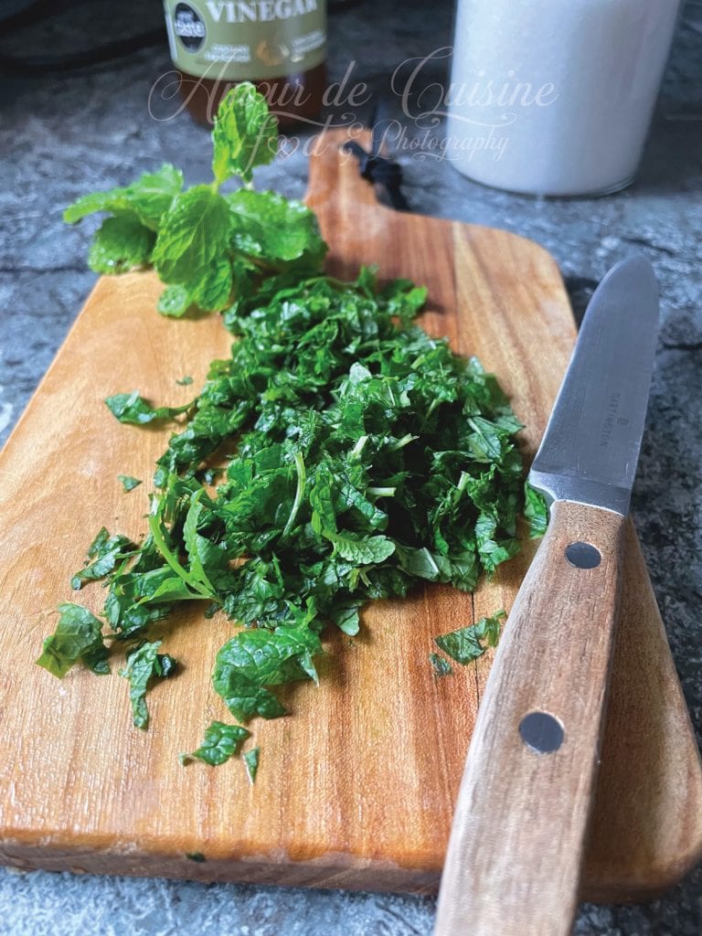 Feuilles de menthe fraîche finement hachées sur une planche en bois, accompagnées d’un couteau à manche en bois. À l’arrière-plan, un bouquet de menthe entière, un pot de sucre et une bouteille de vinaigre posés sur un plan de travail en pierre.