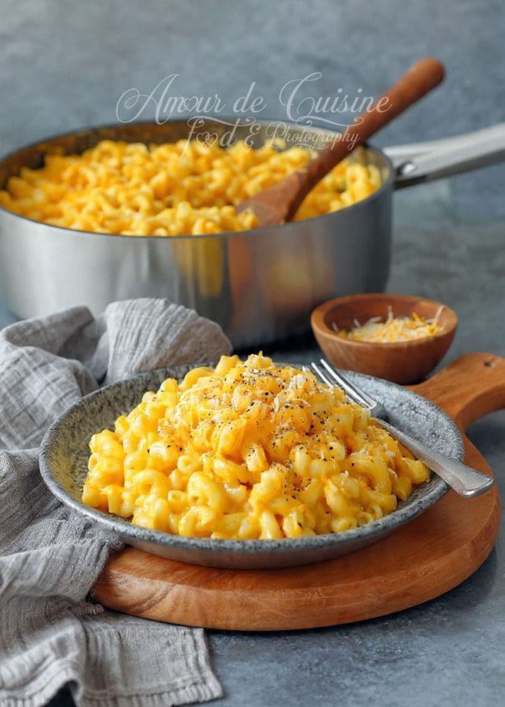 Assiette de mac and cheese crémeux, riche en cheddar et poivre, posée sur une planche en bois, avec une grande casserole remplie du même plat en arrière-plan et un bol de fromage râpé à côté.