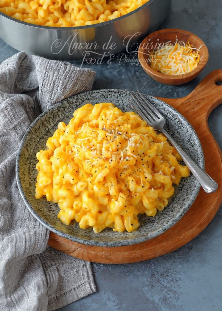 Assiette de mac and cheese crémeux avec macaroni enrobés d’une sauce au cheddar fondante, garnis de poivre et de fromage râpé, posée sur une planche en bois avec une casserole pleine du même plat en arrière-plan.