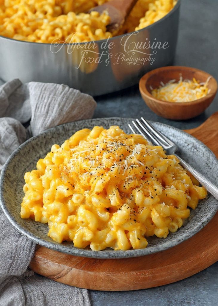 Assiette de mac and cheese crémeux avec macaroni enrobés d’une sauce au cheddar bien fondante, saupoudrés de poivre et de fromage râpé, servie devant une casserole pleine du même plat.