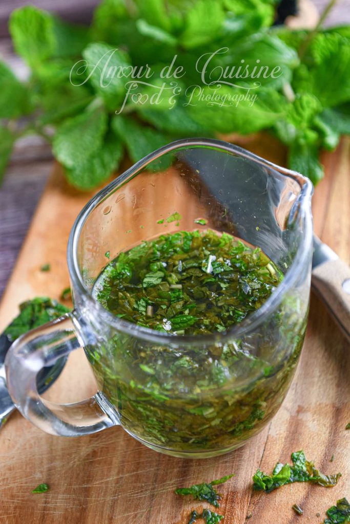 Petit pichet en verre rempli de sauce à la menthe anglaise, composé de feuilles de menthe finement hachées infusées dans un mélange sucré et vinaigré. Le pichet est posé sur une planche en bois, entouré de menthe fraîche et de morceaux de feuilles coupées.