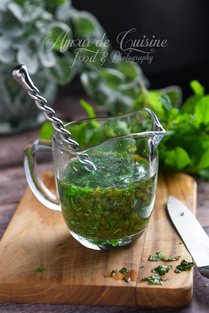 Sauce à la menthe anglaise dans un petit pichet en verre, remplie d’eau infusée et de menthe fraîche finement hachée, avec une cuillère mélangeuse torsadée. Le tout est posé sur une planche en bois, entouré de feuilles de menthe fraîches et d’un couteau.