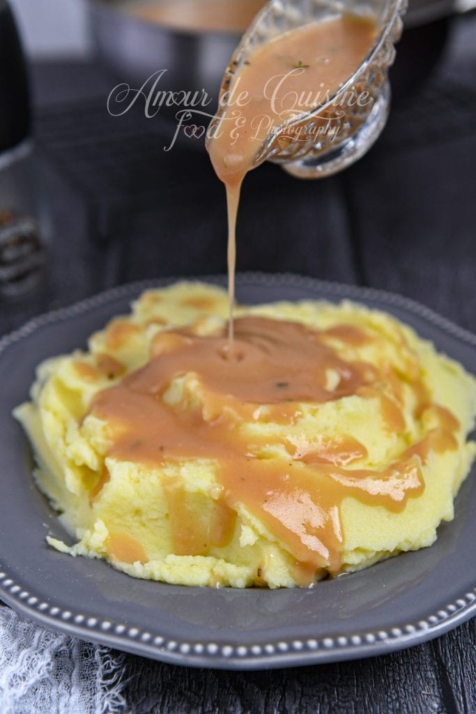 Saucière en verre versant un filet de sauce brune maison sur une assiette de purée de pommes de terre crémeuse. La gravy coule en un ruban lisse et chaud, nappant la purée jaune pâle. En arrière-plan, ambiance sombre et chaleureuse autour de la table.