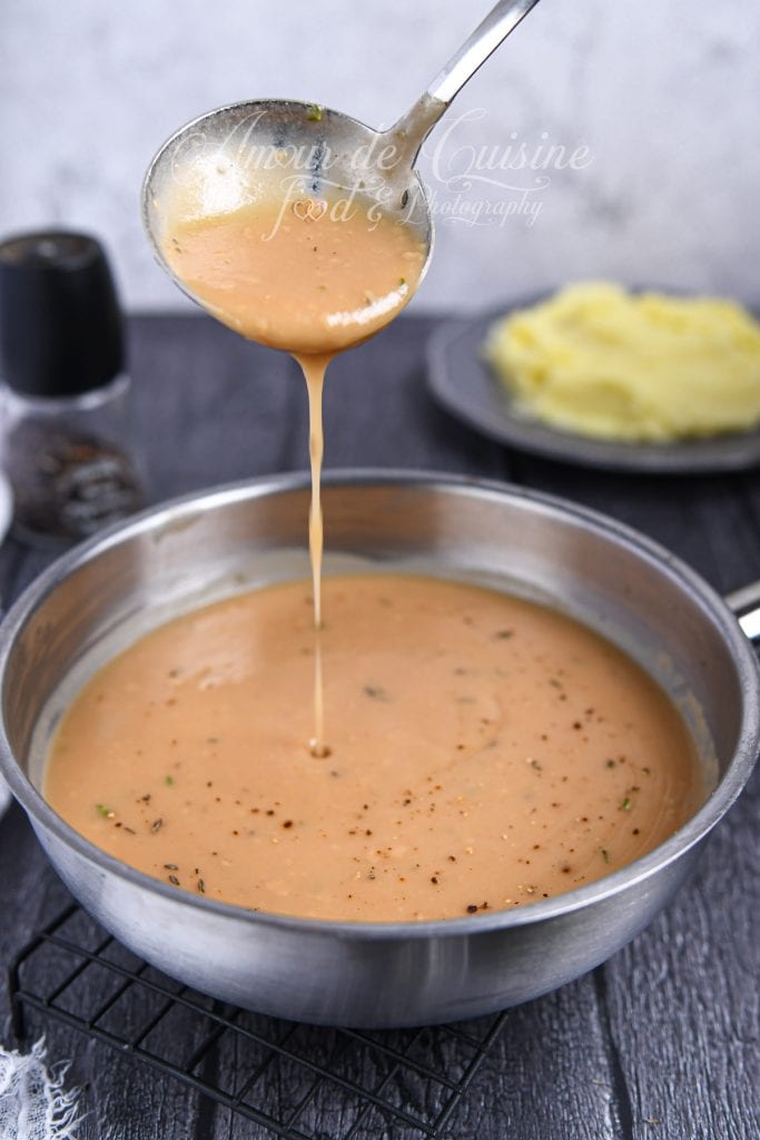 Louche en métal soulevant et versant une sauce brune maison lisse et crémeuse au-dessus d’une grande poêle remplie de gravy, parsemée de poivre noir et d’herbes. En arrière-plan flouté, on aperçoit une assiette de purée et un moulin à poivre sur une table sombre.