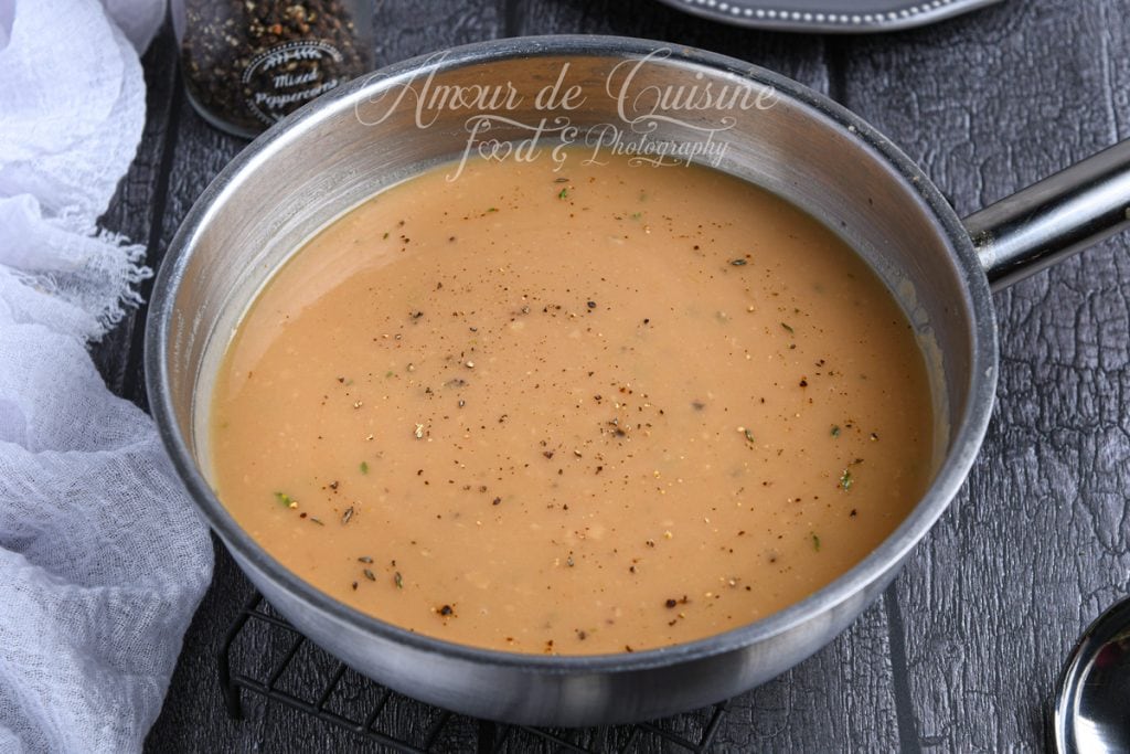 Poêle en inox remplie de sauce brune maison onctueuse, légèrement parsemée de poivre noir et de petites herbes. La sauce a une texture lisse et brillante. En arrière-plan, un moulin à poivre et un tissu blanc reposent sur une surface sombre.