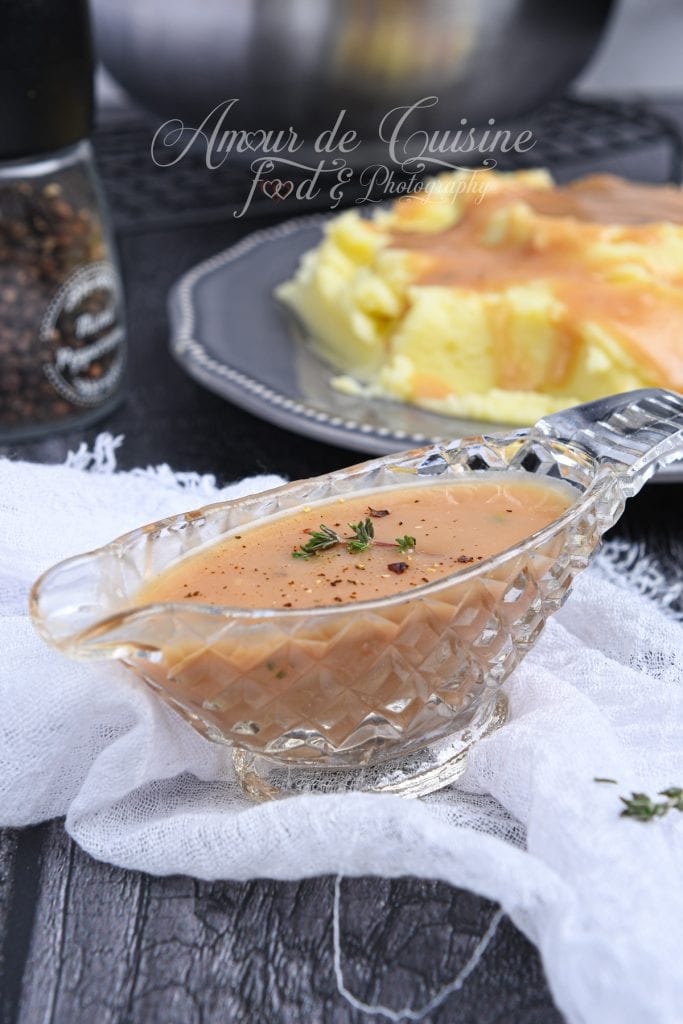 Saucière en verre ciselé remplie de sauce brune maison crémeuse, décorée de poivre noir et de petites feuilles de thym. À l’arrière-plan, une assiette de purée de pommes de terre nappée de gravy est légèrement floutée, posée sur une table sombre avec un moulin à poivre.