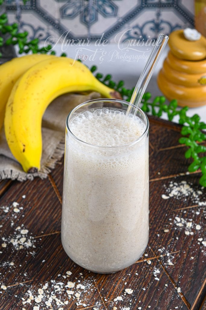 Smoothie banane et avoine crémeux servi dans un grand verre avec une paille transparente, posé sur un plateau en bois, entouré de bananes mûres, de flocons d’avoine et d’un pot de miel en arrière-plan.